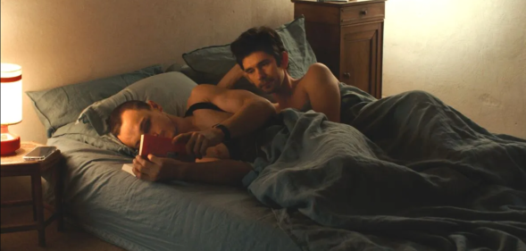 Franz Rogowski as Tomas and Ben Whishaw as Martin in new film Passages. Martin and Tomas are in bed. Tomas is reading, while Martin is looking at him wistfully.