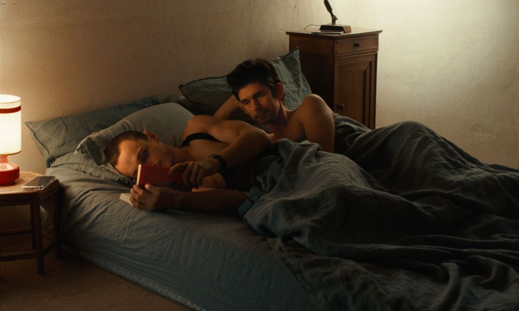 Franz Rogowski as Tomas and Ben Whishaw as Martin in new film Passages. Martin and Tomas are in bed. Tomas is reading, while Martin is looking at him wistfully.