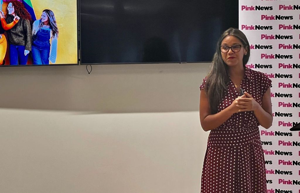 This is an image speaking at an event. She is wearing a read dress with white dots. She is wearing glasses.