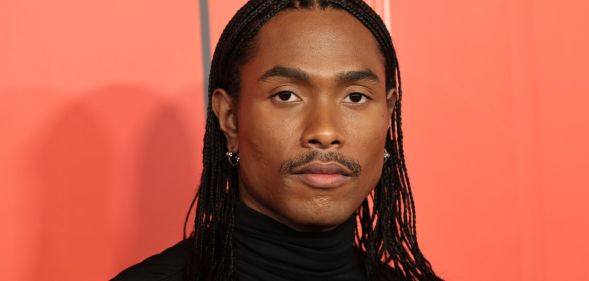 Steve Lacy in a high-necked black top, stood against an orange background.