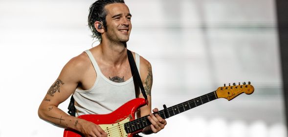 Matty Healy performs on stage in a white vest. He is playing a red electric guitar.