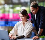 This is an image of two female presenting people at work. One person is sitting at a desk looking at a computer and they are looking at a computer. The other person is standing over them.