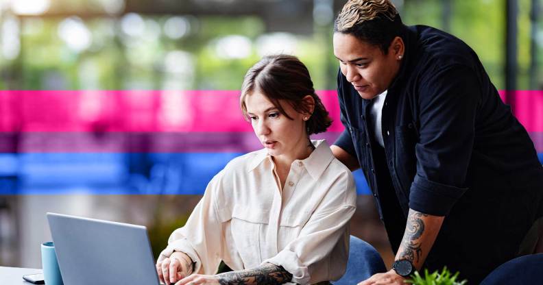 This is an image of two female presenting people at work. One person is sitting at a desk looking at a computer and they are looking at a computer. The other person is standing over them.