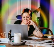 This is an image of a woman working from her home. Her computer is on a table filled with clutter. This imaged is stylised with a rainbow prism effect.