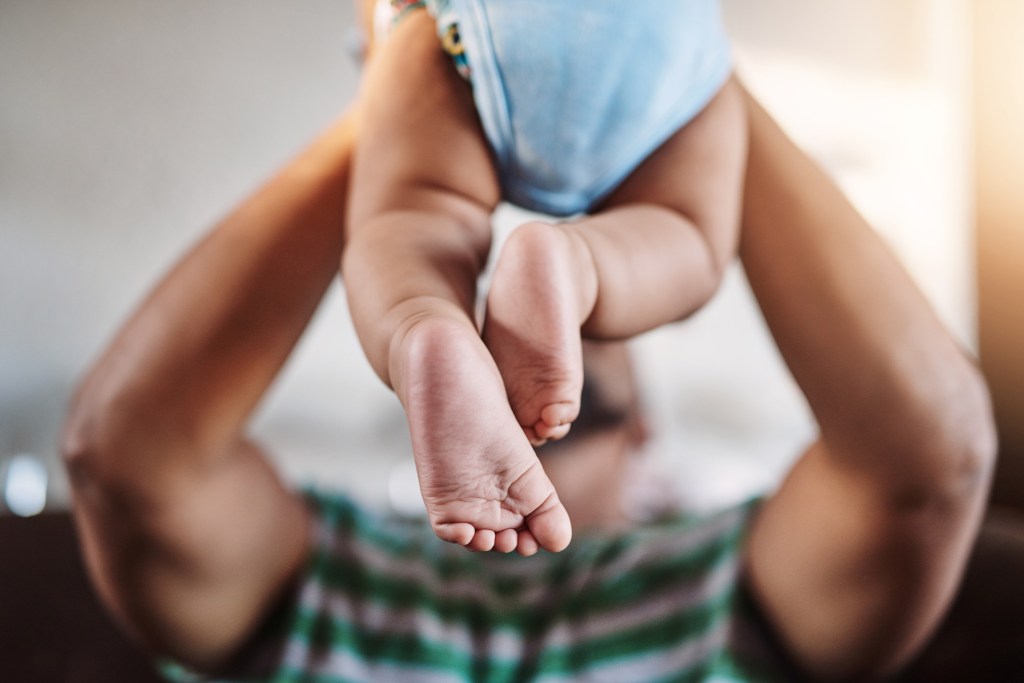 This picture shows a Black man holding his baby up over his head. The man's head is obscured by his baby and all that can be seen is his arms on the left and right hand side of the picture and the baby's legs.