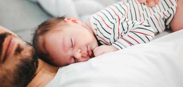 In this image, a man lies on a bed wearing a white t-shirt. His baby lies sound asleep on his chest as he cradles the child.