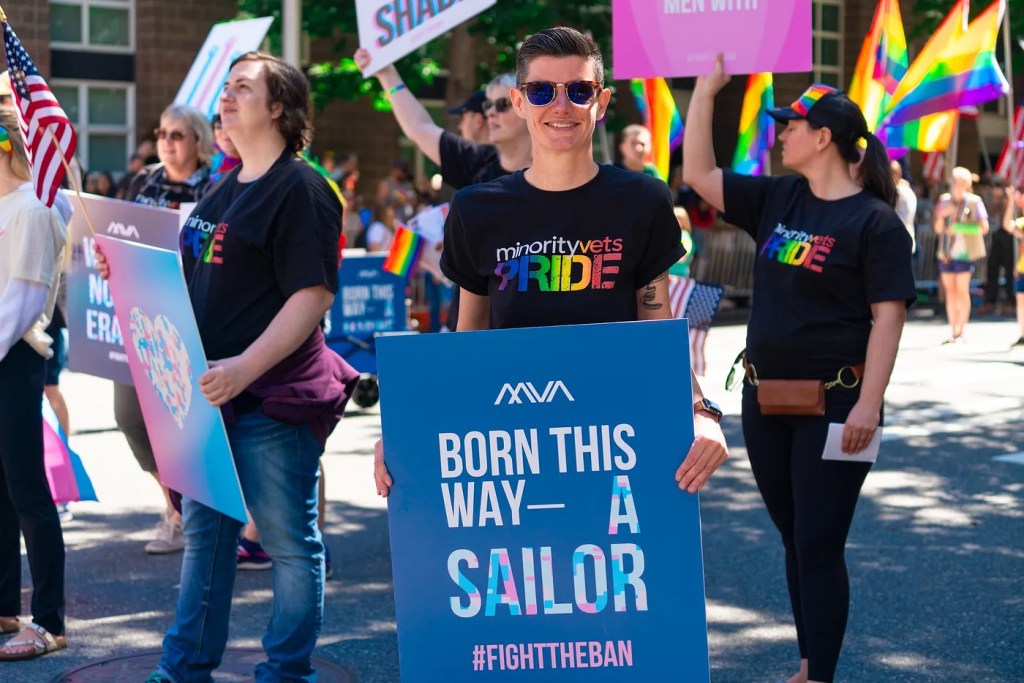 Non-binary Navy veteran and activist Lindsay Church holds up a sign reading 'born this way - a sailor' in protest against anti-LGBTQ+ military policies like Don't Ask, Don't Tell and the trans military ban