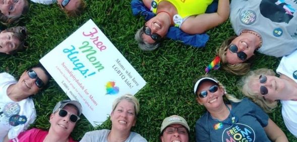 Liz Dyer and a group of moms of LGBTQ+ youth who are part of the Mama Bears group sit on the grass with a 'free hugs' sign