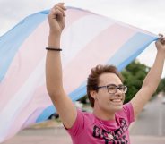 Person wearing a pink shirt and smiling, waving a transgender Pride flag behind them