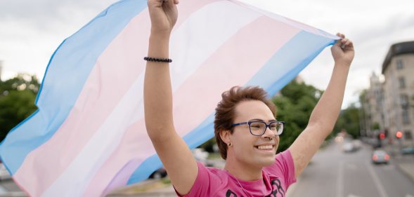 Person wearing a pink shirt and smiling, waving a transgender Pride flag behind them