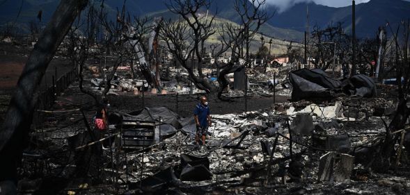 A ground view of the devastation left behind in Maui, Hawaii after wildfires burned down Lahaina. In the picture, there are two people shifting through the rubble, blackened trees and the remains of buildings