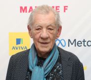 Ian McKellen wears a dark shirt, blue patterned silk scarf and dark grey jacket as he stares towards the camera