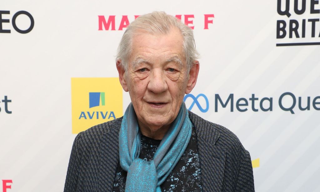 Ian McKellen wears a dark shirt, blue patterned silk scarf and dark grey jacket as he stares towards the camera