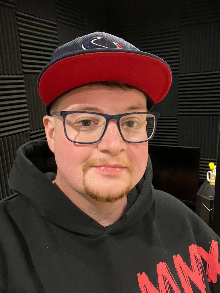 John Junior pictured wearing a cap and a hoodie. He has glasses on and is pictured against a black background.