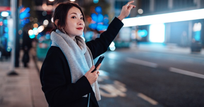 Woman arranging taxi service using mobile app on smart phone, standing on the city street, with illuminated street lights in the background.