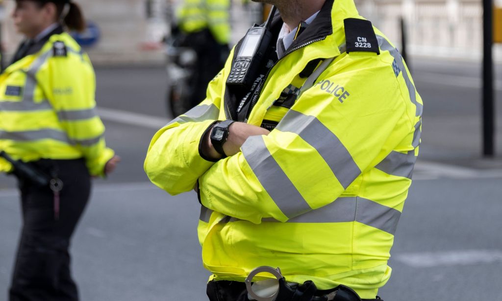 A male Metropolitan Police officer wears yellow reflective gear as he crosses his arms.