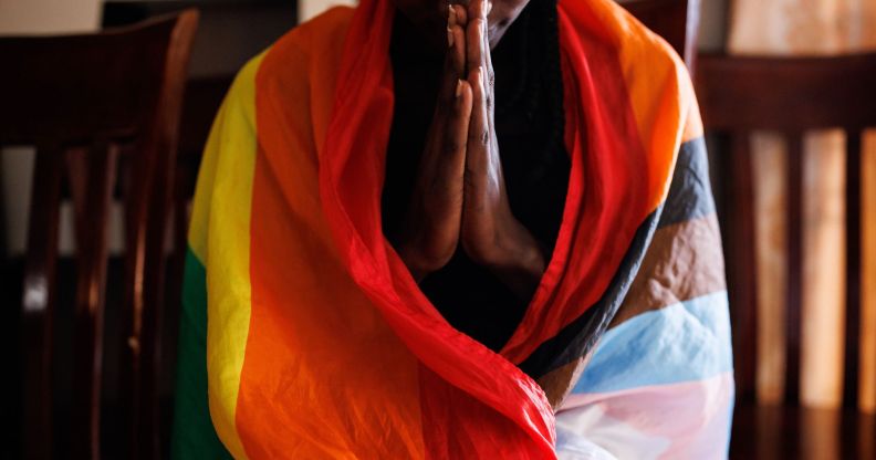 A person in Uganda wears an LGBTQ+ Progressive Pride flag draped around their shoulders as they hold their hands in front of them in prayer