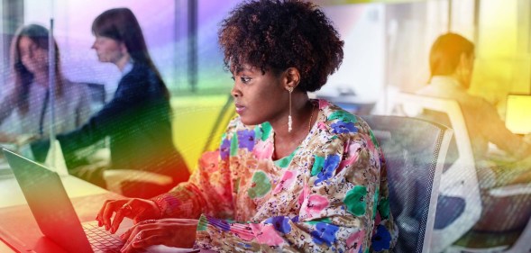 This is an image of a Black woman working at a computer. She is wearing a floral top. There is a creative overlay with the Pride colours over the top