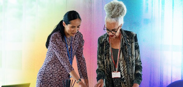 This is an image of two women. The woman on the left is wearing a dress and pointing down at a table. The other woman is wearing a green floral print blazer