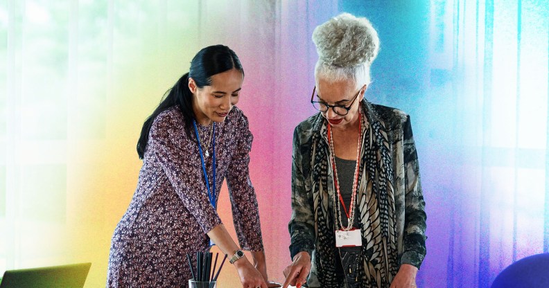 This is an image of two women. The woman on the left is wearing a dress and pointing down at a table. The other woman is wearing a green floral print blazer