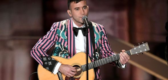 Sufjan Stevens wears a pink and black striped suit jacket as he plays the guitar and sings into a microphone on stage