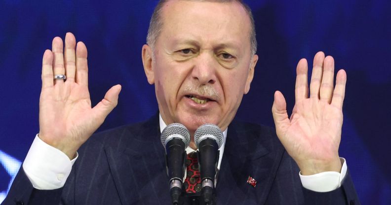 Turkey's president Recep Tayyip Erdoğan, who has a history of making anti-LGBTQ+ remarks, wears a suit and tie as he holds up both his hands while speaking into a microphone