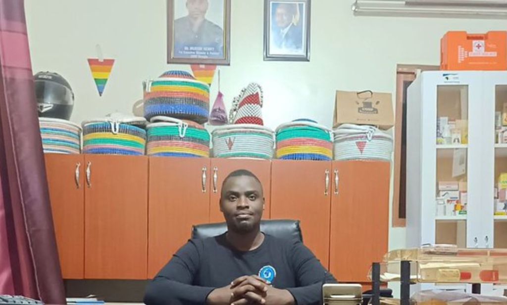 Henry Mukiibi, an LGBTQ+ activist from Uganda, wears a dark shirt as he sits at a desk with several items behind him