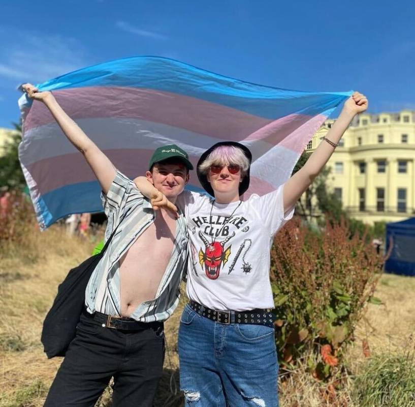 Oli, who has top surgery, and his non-binary sibling hold up a trans Pride flag while standing on a Brighton beach