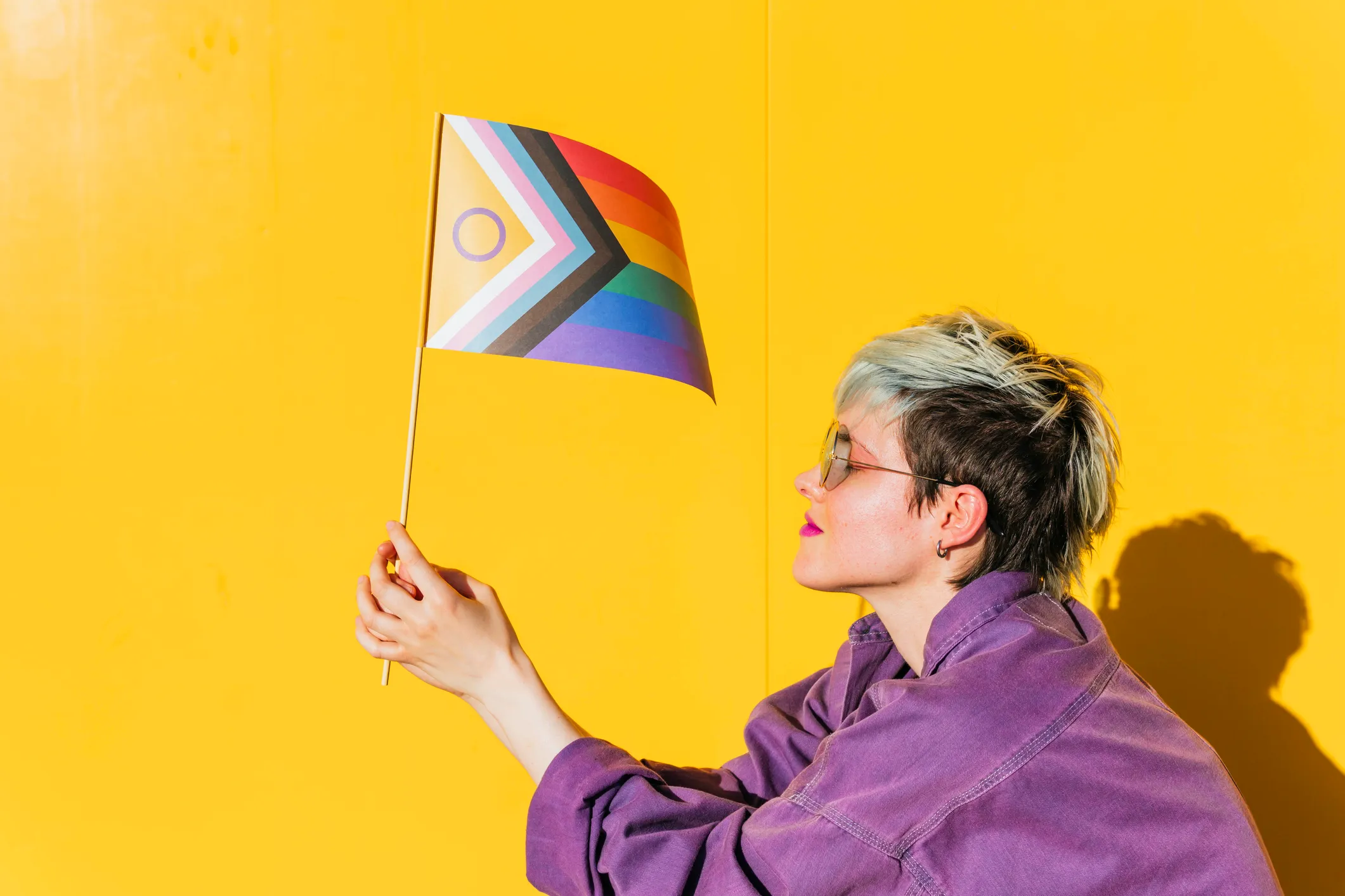 Lesbian woman holding multi colored flag and squatting in front of yellow wall